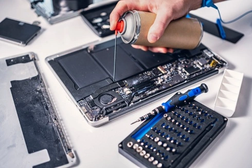The image shows a person using compressed air to clean the internal components of a laptop.