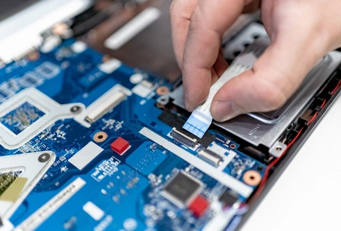 The image shows a technician carefully working on a laptop's motherboard, specifically handling a ribbon cable that connects internal components