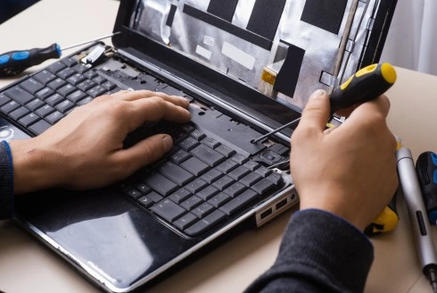 The image shows a person working on a laptop, using a screwdriver to carefully disassemble the device.
