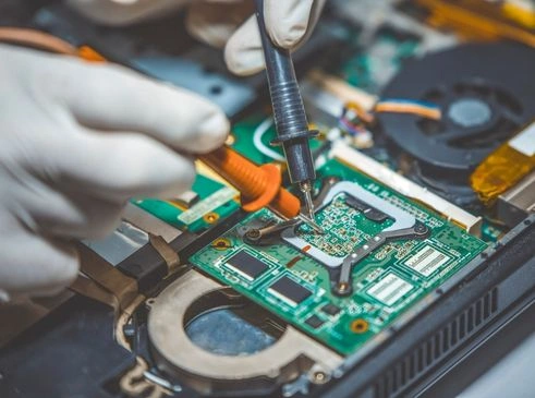 The image shows a technician wearing gloves and using a soldering iron to work on a laptop's motherboard