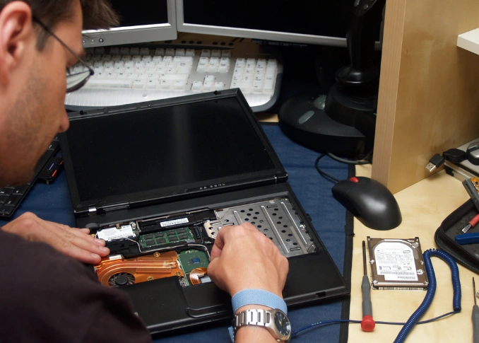 A technician repairing a laptop on a desk, with the screen open and internal components exposed, surrounded by multiple monitors and tools.